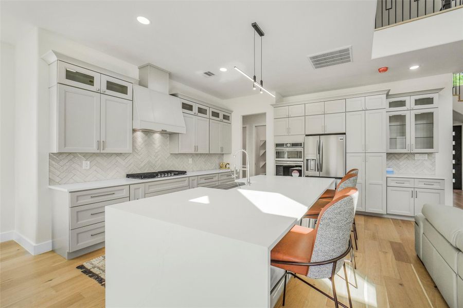 This modern kitchen features sleek white cabinetry, a spacious island with seating, and stainless steel appliances. It has a bright, open layout with light wood flooring and contemporary lighting, perfect for entertaining.