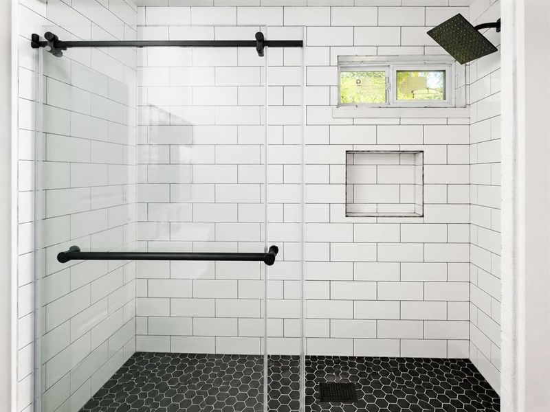 Modern shower enclosure featuring clear glass panels with matte black hardware, white subway tile walls, a hexagonal mosaic tile floor, an integrated shower niche, and a fixed rainfall showerhead