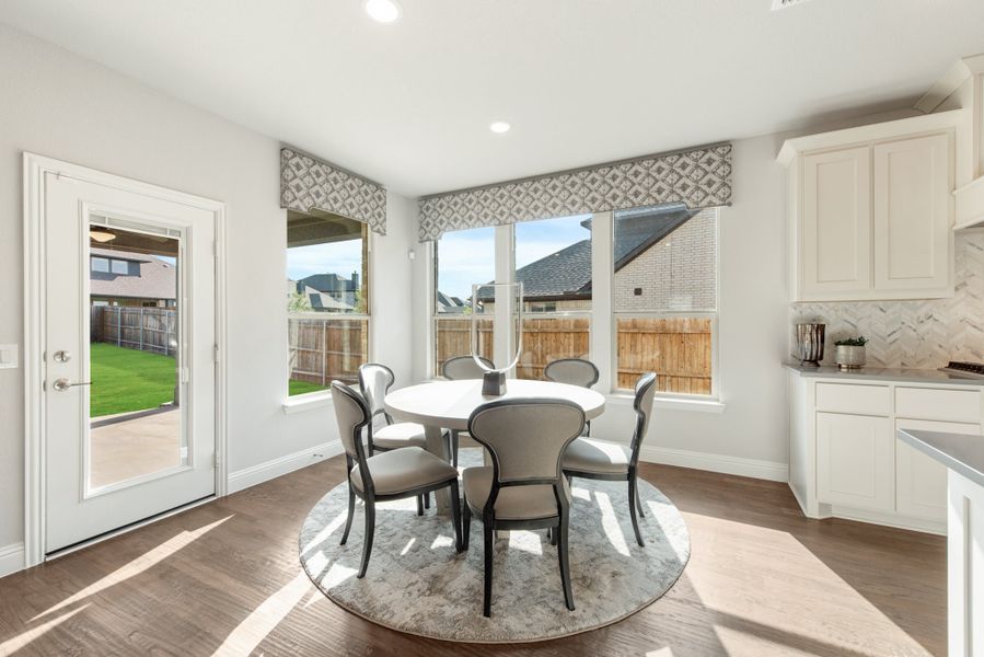 Breakfast nook with round white table, six gray chairs, hardwood floors, and large windows overlooking backyard