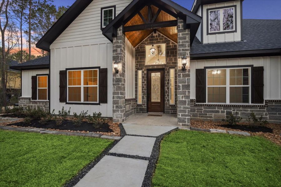 Close view of exterior entry to the home.  Full light front door with side lights brings extra natural light into the home.  Square picture window above the door frames the indoor light fixture and adds interest to the home exterior.   Exterior also has hanging light and two coach lights. Stained wood above the covered entry.  The window on the right side of the entry is the home office. Upper windows are decorative.  The windows on the left side of the home are secondary bedrooms.