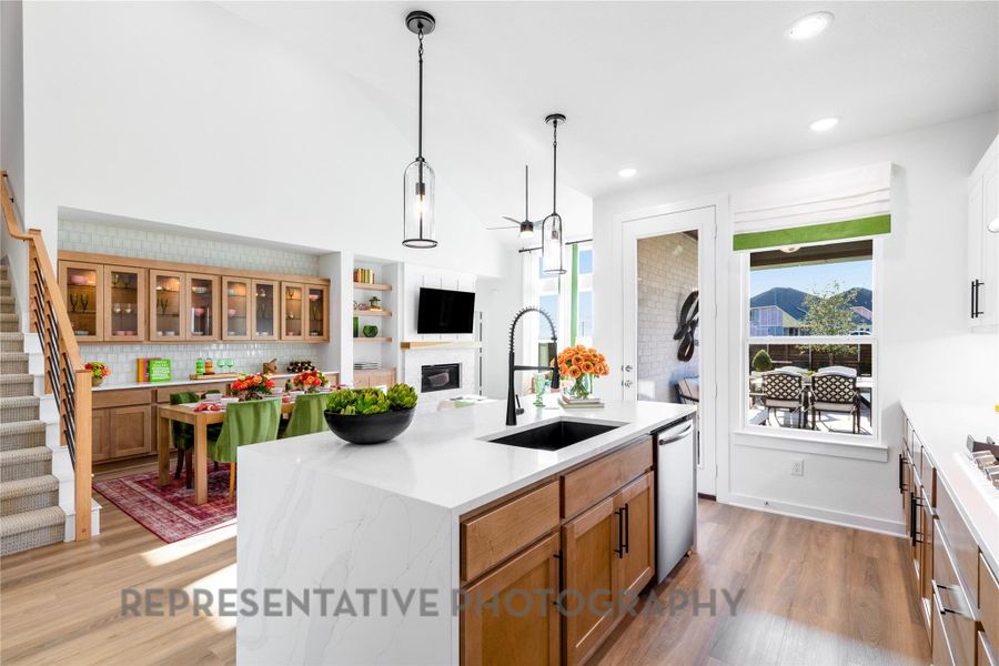 Kitchen with dishwasher, brown cabinetry, light wood-type flooring, a center island with sink, and a glass covered fireplace