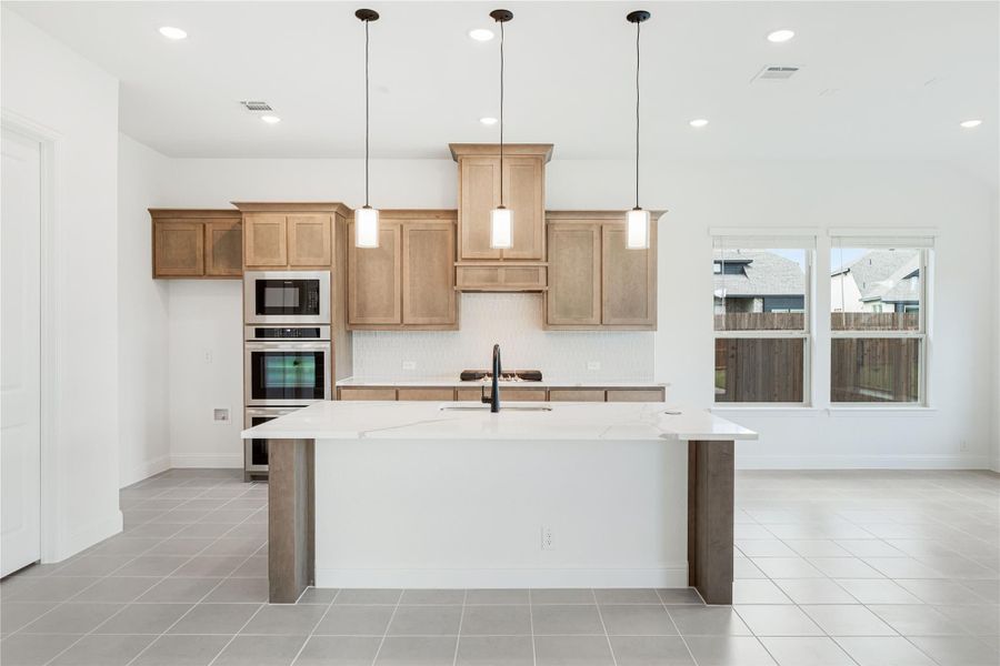 Kitchen with large island, light wood cabinets, double oven, and three pendant lights over white countertops