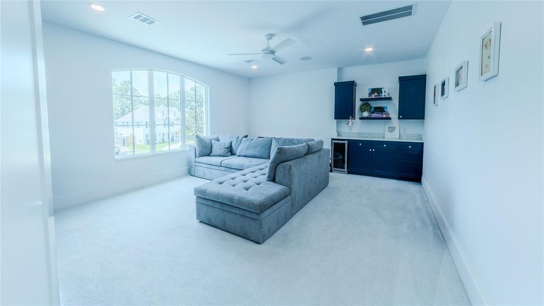 This room offers a bright and inviting space with a large window, plush gray sectional sofa, and cozy carpet. It features built-in dark cabinetry with shelves and a small fridge, perfect for relaxation and entertainment.