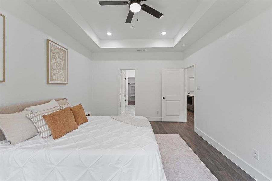 Spacious bedroom featuring a tray ceiling with recessed lighting and a ceiling fan, wood-finish flooring, and white interior doors with dark hardware Spacious bedroom featuring a tray ceiling with recessed lighting and a ceiling fan, wood-finish flooring, and white interior doors with dark hardware