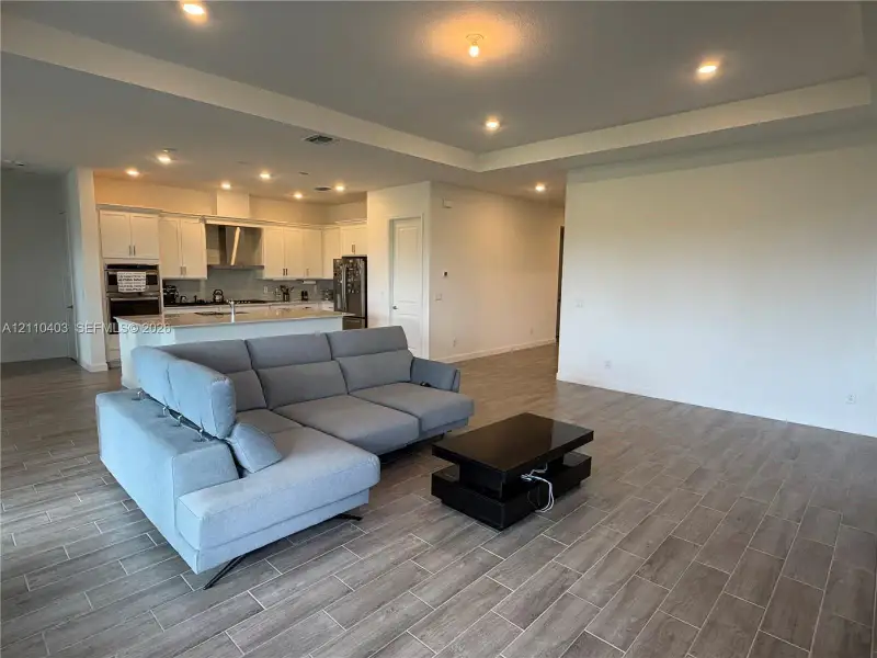 Open-concept living space featuring wood-finish tile flooring, recessed lighting, tray ceilings, and a kitchen equipped with white cabinetry and a central island