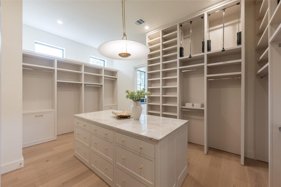 The expansive primary closet features custom millwork with double pull-down rods, a built-in island with quartzite counters, and brass hardware. Natural light from clerestory windows enhances the serene, boutique-like atmosphere. The expansive primary closet features custom millwork with double pull-down rods, a built-in island with quartzite counters, and brass hardware. Natural light from clerestory windows enhances the serene, boutique-like atmosphere.