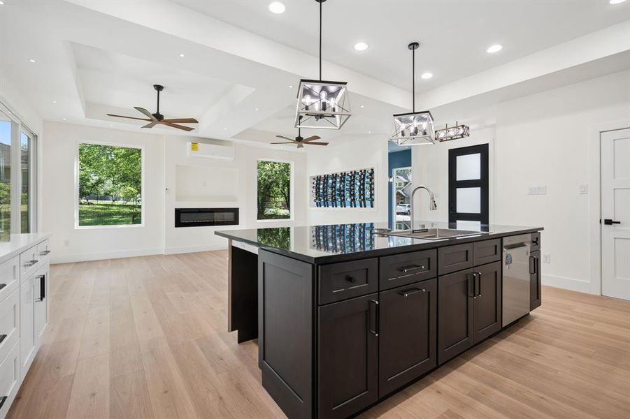 Two tone kitchen with a kitchen island with sink, a glass covered fireplace, light wood finished floors, stainless steel dishwasher, and two tone cabinets. Space for two undercabinet refrigerators.