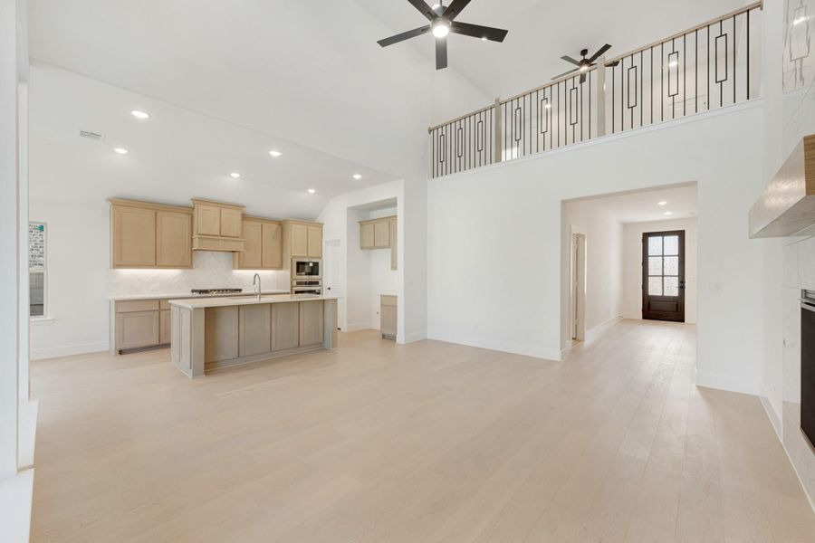 Open-concept living area with kitchen island, light wood floors, high vaulted ceiling, and second-floor balcony overlook