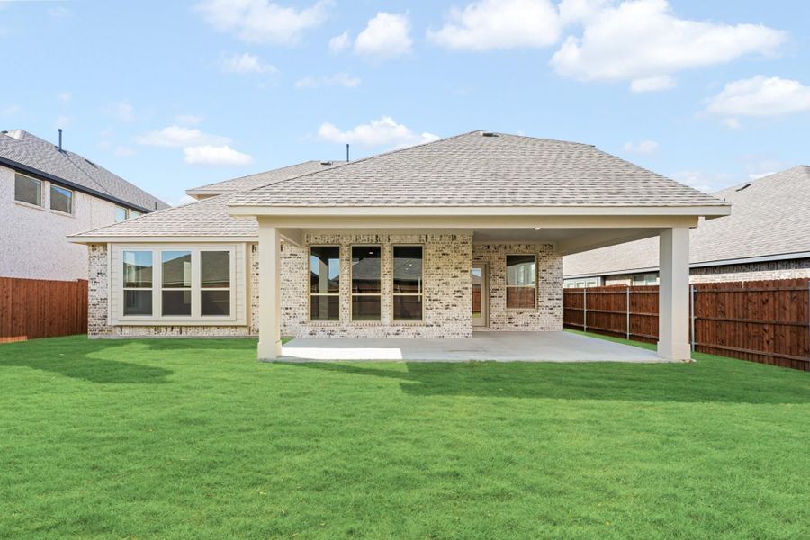 Rear view of brick home with covered patio, concrete slab, green lawn, and wood privacy fence.