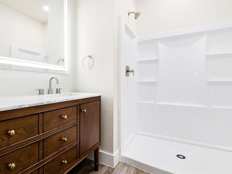 Bathroom featuring a wood-finish vanity with a stone countertop, an illuminated mirror, a brushed nickel faucet, and a white shower stall with a brushed nickel shower head Bathroom featuring a wood-finish vanity with a stone countertop, an illuminated mirror, a brushed nickel faucet, and a white shower stall with a brushed nickel shower head