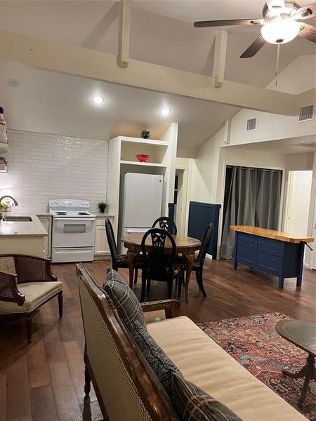 Open-concept living area featuring vaulted ceilings with exposed beams, wood-finish flooring, recessed lighting, and a kitchen with white subway tile backsplash and granite-finish countertops
