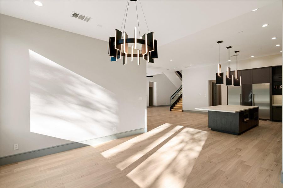 This modern open-concept living space features sleek lighting fixtures, light wood flooring, and ample natural light. The kitchen includes a large island and stainless steel appliances, while the area leads to a staircase with a contemporary design.