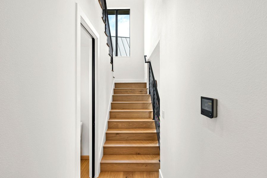 A modern wooden staircase rises past white walls toward a tall upper window.