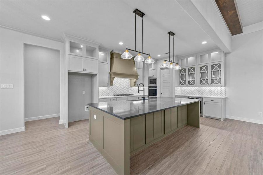 Kitchen featuring custom exhaust hood, hanging light fixtures, light wood-type flooring, an island with sink, and tasteful backsplash