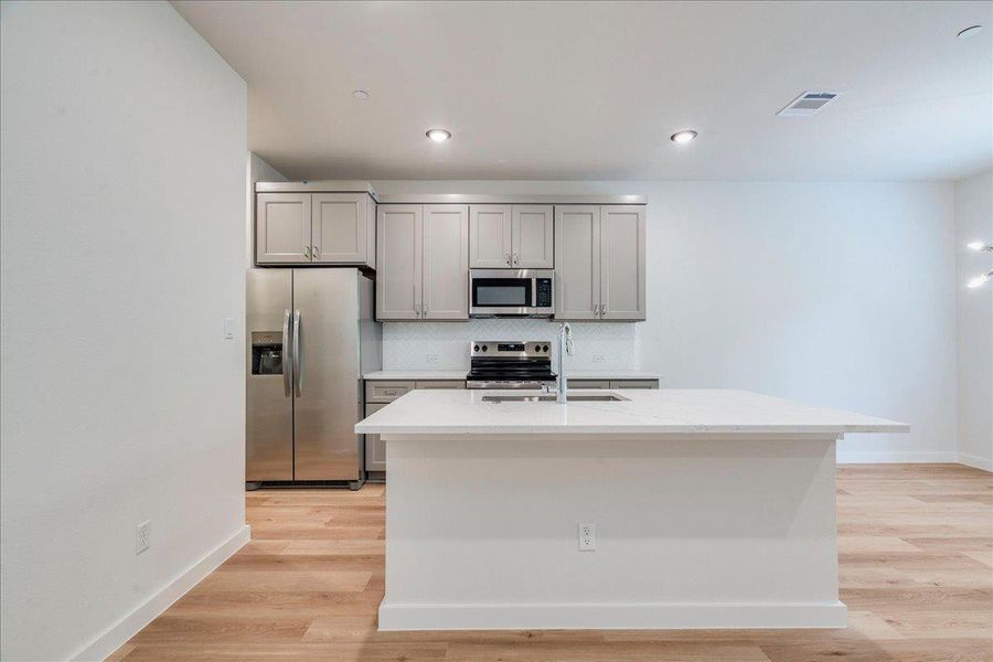 A sleek island anchors this well-lit kitchen, boasting contemporary appliances and light wood flooring.