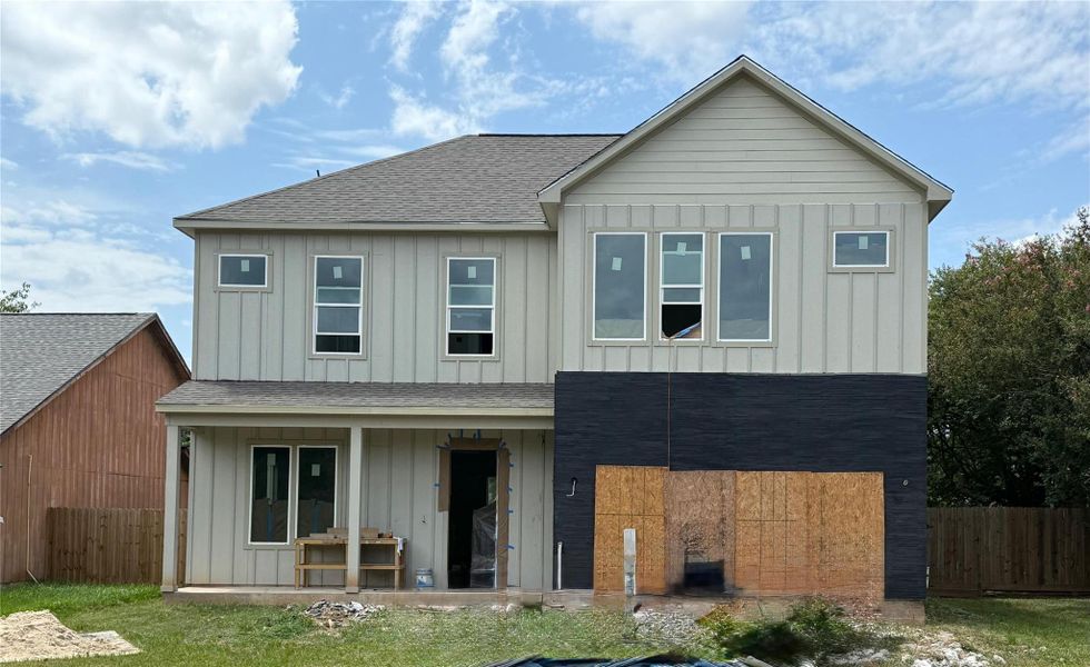 This photo shows a two-story house under construction with a modern design. The exterior features vertical siding, a covered porch, and a partially completed facade.