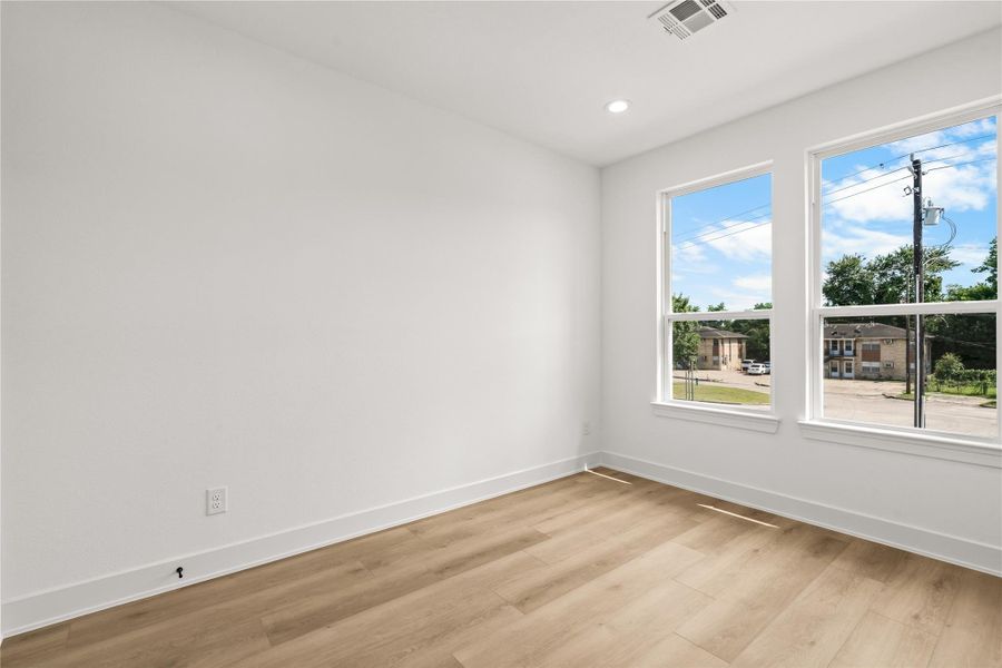 The second bedroom features a bright, airy layout with white walls and warm flooring. Two large vertical windows provide plenty of natural light and neighborhood views, while the spacious layout easily fits a queen-sized bed and extra storage furniture.