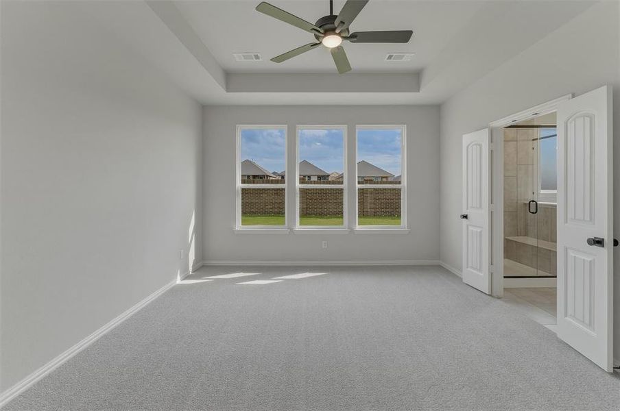 Spacious room featuring light gray carpeting, a tray ceiling with a dark-bladed ceiling fan, and three large windows
