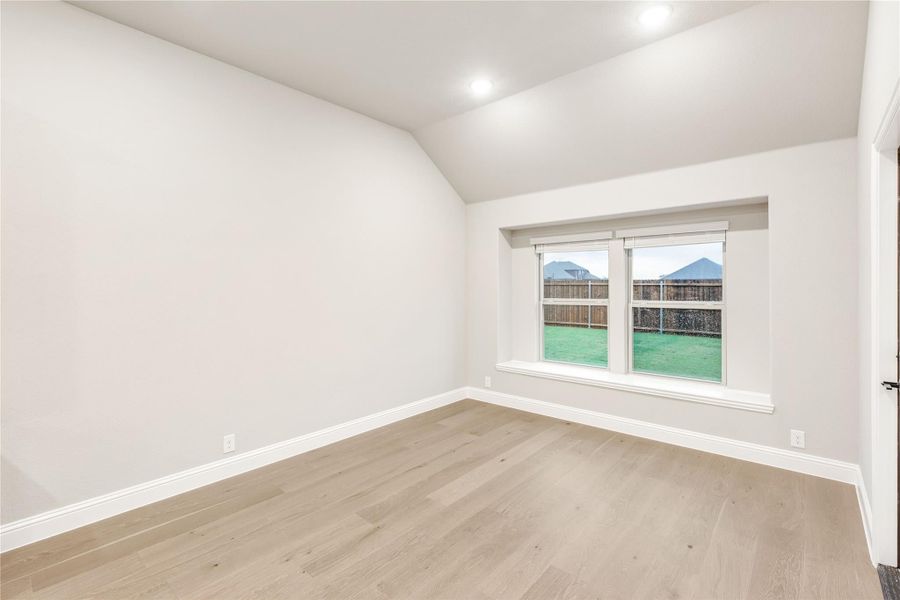 Empty room with vaulted ceiling, light hardwood floors, and a window overlooking a fenced backyard
