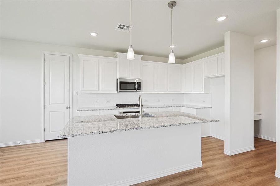 Kitchen featuring light wood-type flooring, an island with sink, decorative light fixtures, white cabinets, and light stone counters