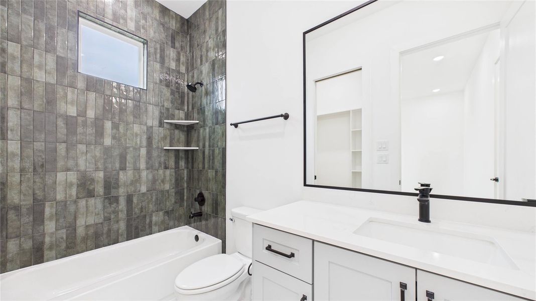 This modern bathroom features sleek, dark tiled walls that reach to the ceiling, with a built-in bathtub and shower. It has a large mirror above a white vanity with black fixtures, providing a clean and stylish look.