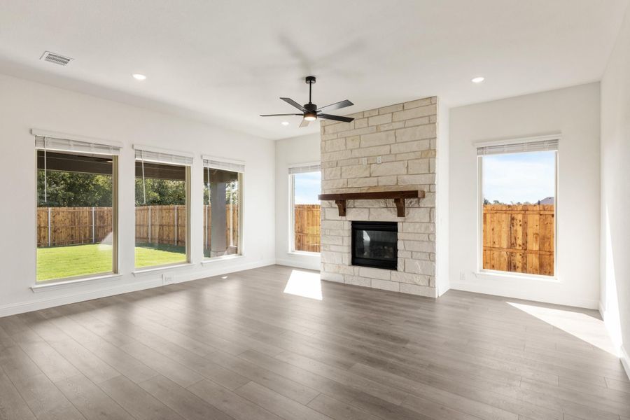 Open living room with stone fireplace, hardwood floors, ceiling fan, and large windows overlooking backyard Open living room with stone fireplace, hardwood floors, ceiling fan, and large windows overlooking backyard