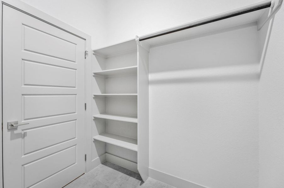 This photo shows a clean, white walk-in closet with built-in shelves and a clothing rod, offering ample storage space. The closet features a modern door with a sleek handle. This photo shows a clean, white walk-in closet with built-in shelves and a clothing rod, offering ample storage space. The closet features a modern door with a sleek handle.