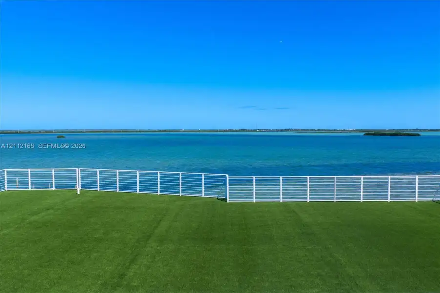 Outdoor space featuring synthetic turf and a white horizontal-slat metal railing, set against a waterfront backdrop Outdoor space featuring synthetic turf and a white horizontal-slat metal railing, set against a waterfront backdrop