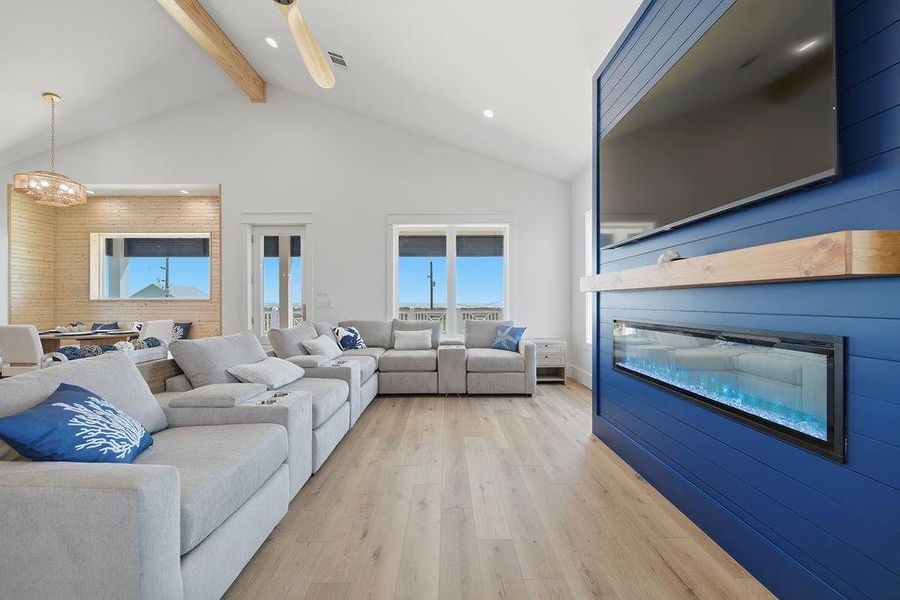 This spacious living room features a modern design with vaulted ceilings, light wood flooring, and a striking blue accent wall with a mounted TV and fireplace. Comfortable gray sectional seating complements the airy, open layout, leading into a dining area with stylish lighting. Large windows provide ample natural light.