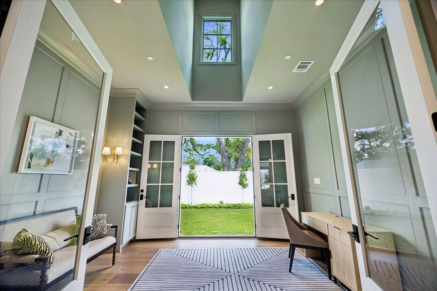 Framed by glass divided light double doors, this impressive study features soaring ceiling with a dramatic ceiling detail and overhead window, allowing natural light to pour in from above. Paneled walls, custom built-ins, and elegant sconces add architectural interest, while French doors open directly to the side yard, creating a seamless indoor-outdoor connection.