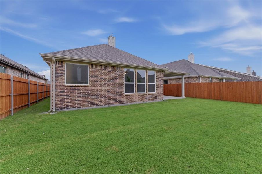 Expansive green lawn with a brown privacy fence, brick exterior, multiple windows, and a covered patio area