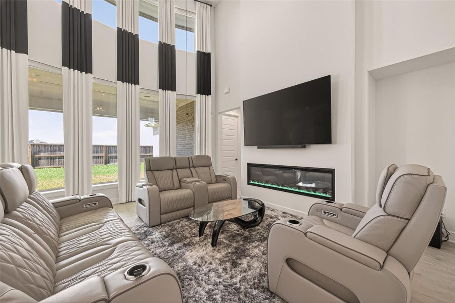 This spacious, modern living room features high ceilings and large windows with elegant curtains, allowing ample natural light. It includes plush reclining sofas, a large wall-mounted TV, and a sleek electric fireplace, creating a cozy, luxurious atmosphere.