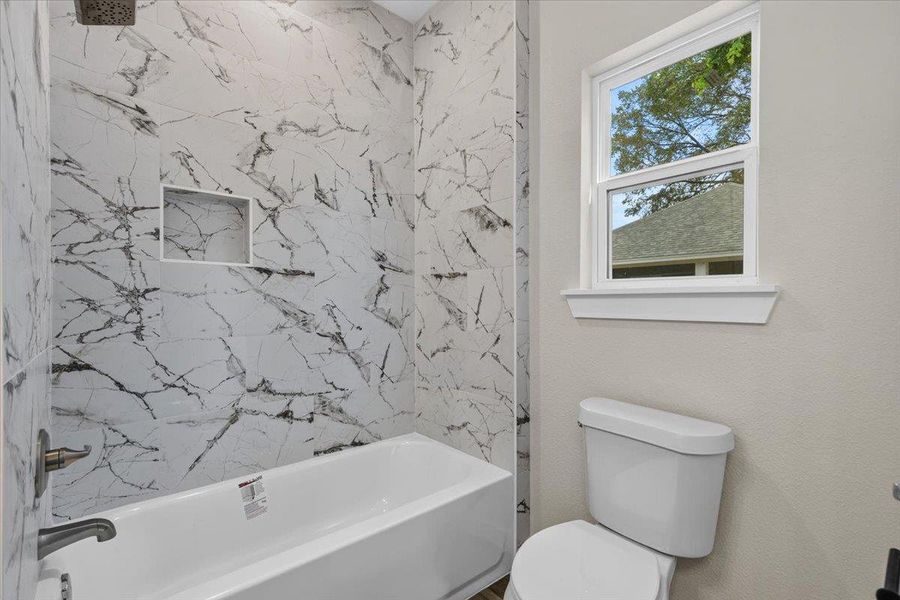 A window overlooks the backyard from this bathroom featuring a marble-patterned tile tub surround. A window overlooks the backyard from this bathroom featuring a marble-patterned tile tub surround.