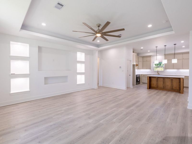 This spacious, open-concept living area features light hardwood floors, recessed lighting, and a modern ceiling fan. The adjacent kitchen boasts a large island with pendant lights, ample cabinetry, and stainless steel appliances, creating a bright and inviting space perfect for entertaining.