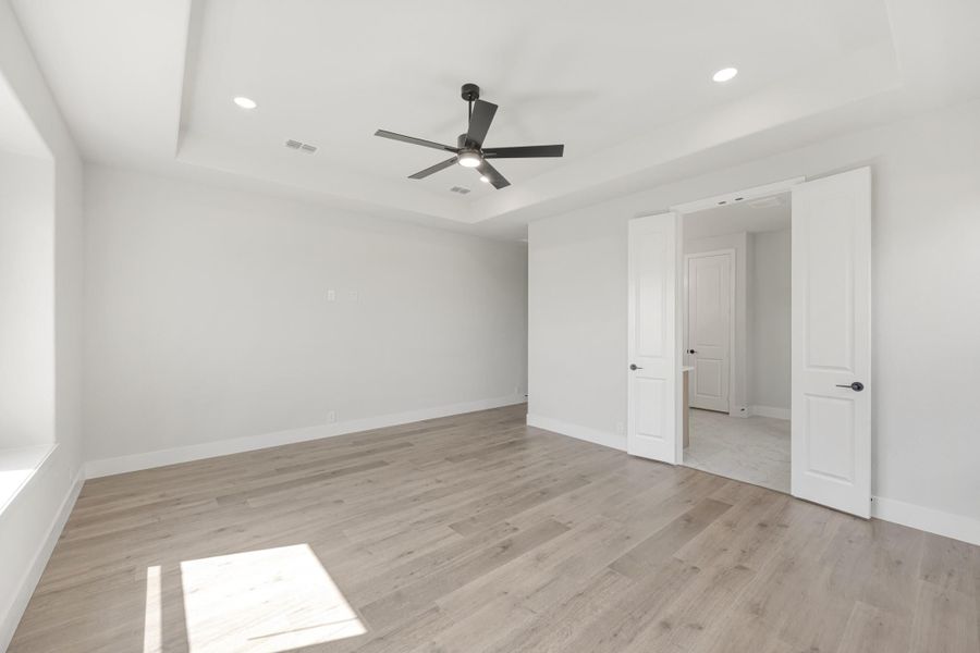 Empty bedroom with tray ceiling, black ceiling fan, light hardwood floors, and white double doors leading to ensuite Empty bedroom with tray ceiling, black ceiling fan, light hardwood floors, and white double doors leading to ensuite