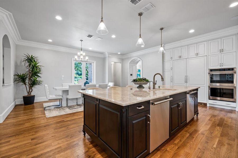 Kitchen featuring appliances with stainless steel finishes, wood-type flooring, light stone countertops, pendant lighting, and a center island with sink