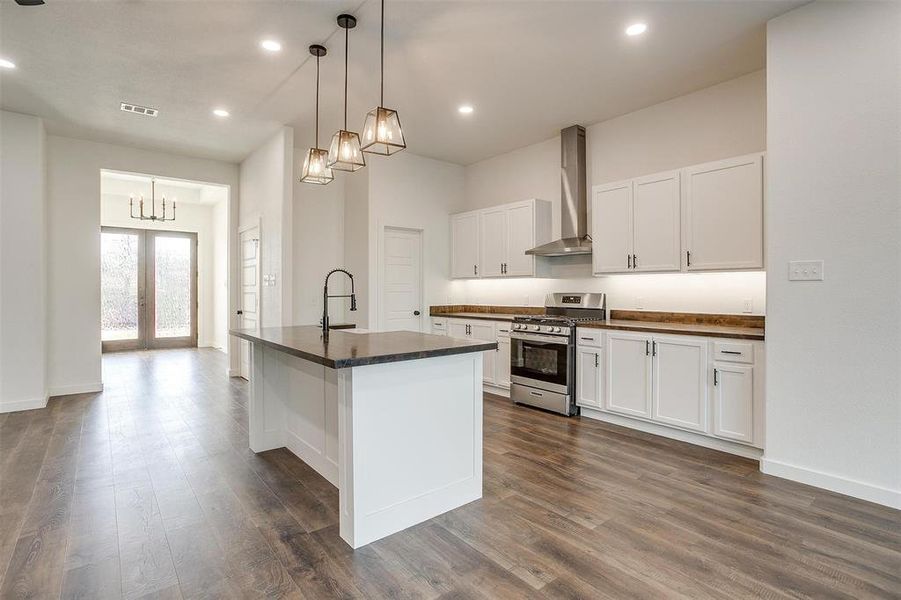 Kitchen featuring a kitchen island with sink, hanging light fixtures, white cabinets, wall chimney range hood, and gas range