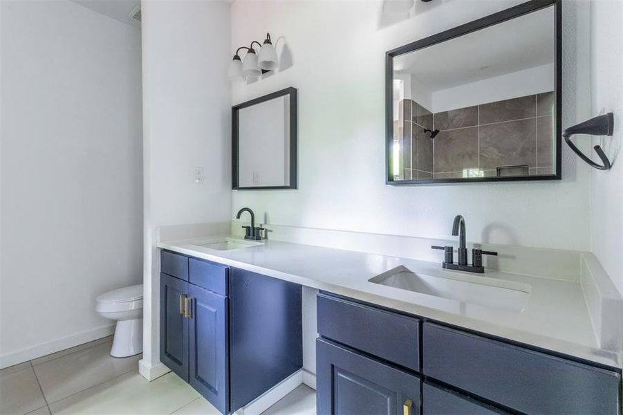 Bathroom with toilet, tile patterned flooring, double vanity, a shower, and baseboards Bathroom with toilet, tile patterned flooring, double vanity, a shower, and baseboards