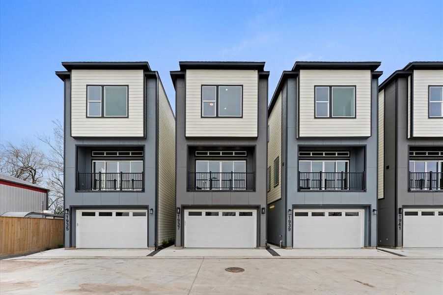 This photo shows a row of modern, three-story townhouses with a sleek design. Each unit features a garage, a small balcony on the second floor, and large windows for natural light. The exterior is a mix of gray and white tones, offering a contemporary look.