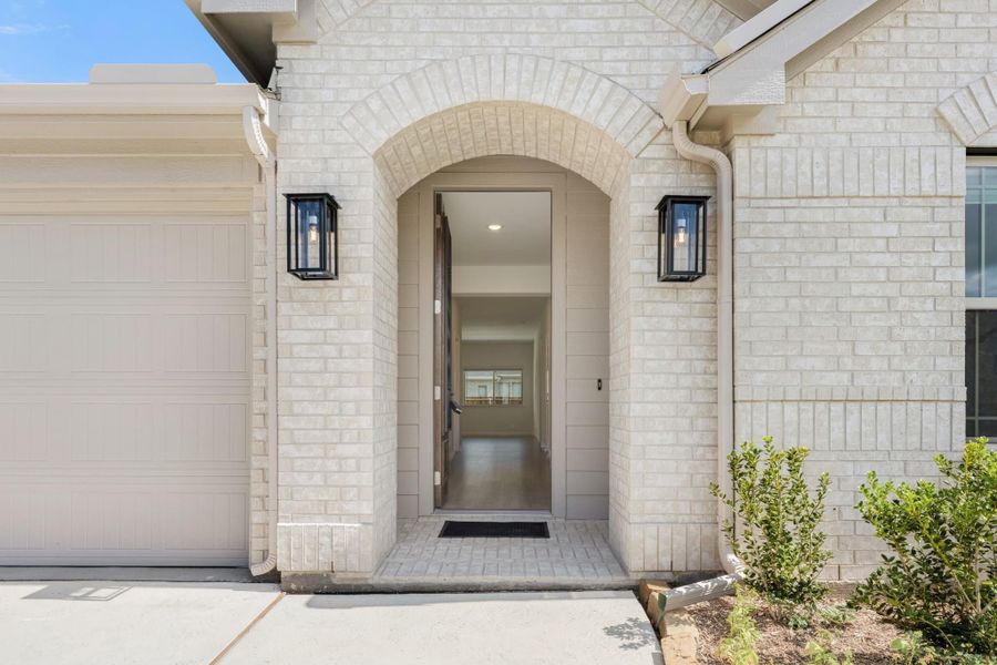 Front entry with arched brick surround, open door, and two black wall sconce lights flanking the entrance Front entry with arched brick surround, open door, and two black wall sconce lights flanking the entrance
