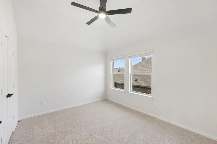 Empty bedroom with beige carpet, ceiling fan, and two windows with neighborhood view