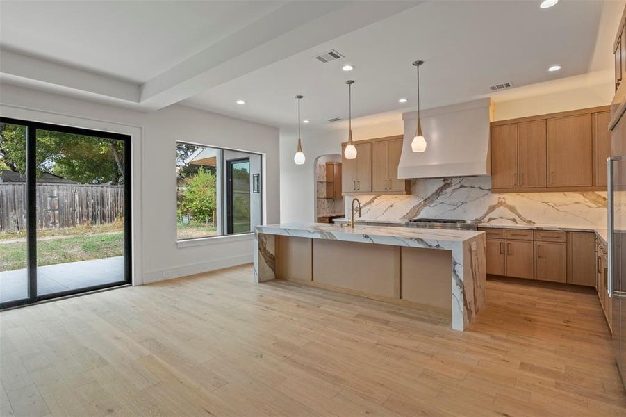 Kitchen featuring light stone counters, backsplash, custom exhaust hood, hanging light fixtures, and a center island with sink Kitchen featuring light stone counters, backsplash, custom exhaust hood, hanging light fixtures, and a center island with sink