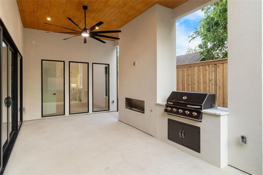 Covered patio featuring a wood-paneled ceiling with a modern ceiling fan, an outdoor kitchen with a built-in grill, stone countertop, and integrated storage, and large glass panel doors Covered patio featuring a wood-paneled ceiling with a modern ceiling fan, an outdoor kitchen with a built-in grill, stone countertop, and integrated storage, and large glass panel doors