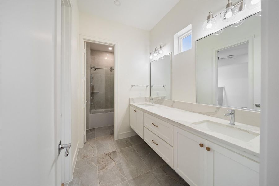 Secondary Jack and Jill bathroom features a modern double vanity with sleek faucets, ample storage, and elegant marble countertops. It has a clean, bright design with a large mirror and a bathtub with a glass door, complemented by stylish lighting fixtures.