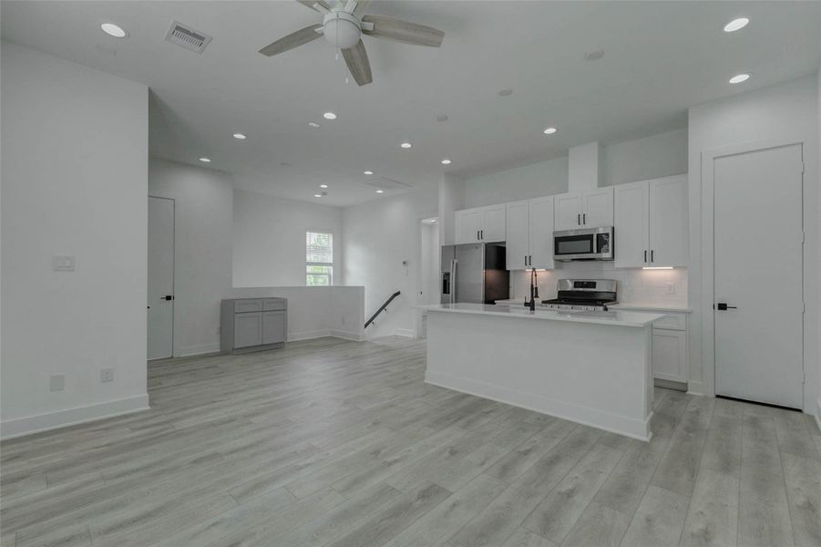 Expansive kitchen and living view featuring a large white island, stainless appliances, white cabinetry, black hardware, under-cabinet lighting, and light wood-look flooring. Tall ceilings, recessed lights, and an open layout give the space a polished contemporary feel with room for dining, gathering, and comfortable daily living.