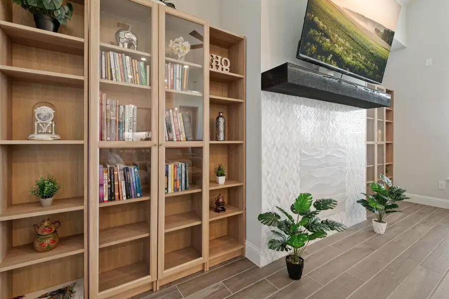 This cozy living space features a stylish built-in bookcase with glass doors, a modern white textured fireplace, and a wall-mounted TV. The wood-look tile flooring and green plants add warmth and freshness to the room. This cozy living space features a stylish built-in bookcase with glass doors, a modern white textured fireplace, and a wall-mounted TV. The wood-look tile flooring and green plants add warmth and freshness to the room.