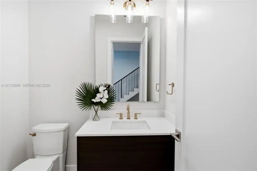 Bathroom featuring a dark wood-finish vanity, white countertop with an integrated basin, wall-mounted rectangular mirror, three-light fixture with cylindrical glass shades, and gold-tone hardware