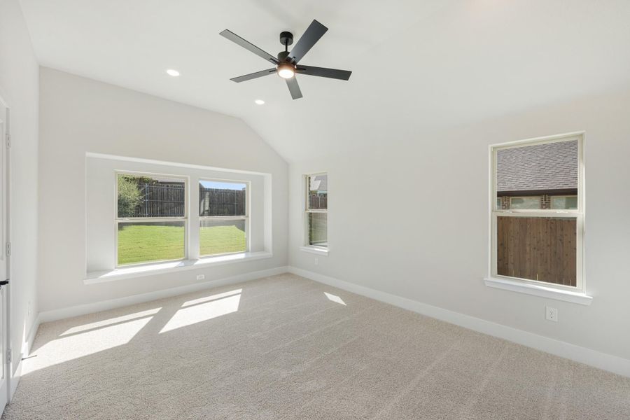 Empty bedroom with vaulted ceiling, ceiling fan, carpet flooring, and multiple windows overlooking backyard Empty bedroom with vaulted ceiling, ceiling fan, carpet flooring, and multiple windows overlooking backyard