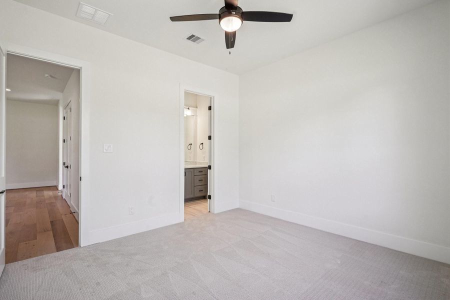 Bright, neutral-toned bedroom with carpeted floor, ceiling fan, and an en-suite bathroom. Hardwood hallway leads to adjacent rooms.