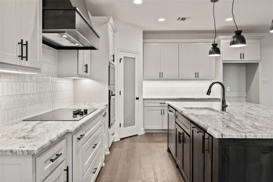 Kitchen with custom exhaust hood, white cabinets, light wood-style floors, light stone counters, and a kitchen island with sink